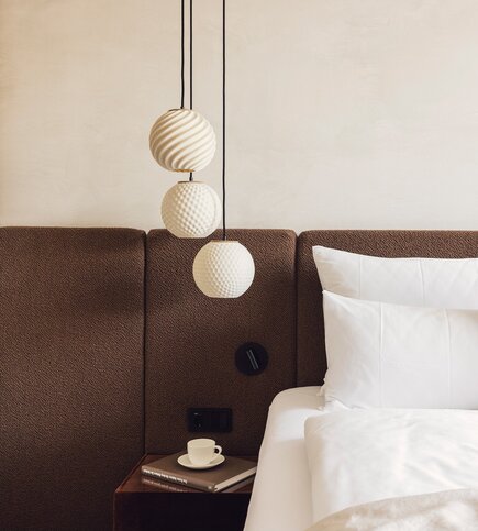 Ein Bett mit weißen Kissen steht neben einem Nachttisch mit Tasse und Buch unter drei Design-Pendelleuchten.