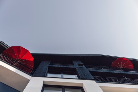Die Ansicht zeigt die Fassade eines Apartmentgebäudes mit Balkonen und zwei roten Sonnenschirmen.
