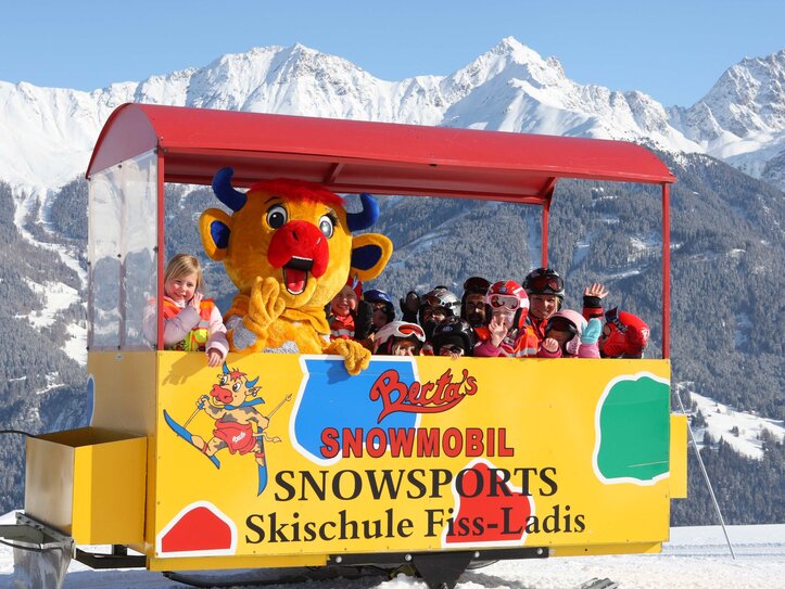 Skischule Fiss-Ladis mit Kindern im Schneewagen Kinder sitzen in einem offenen Schneewagen vor einer Skischule in Fiss-Ladis, begleitet von einer bunten Maskottchenfigur.
