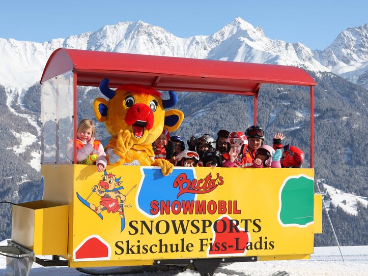 Skischule Fiss-Ladis mit Kindern im Schneewagen Kinder sitzen in einem offenen Schneewagen vor einer Skischule in Fiss-Ladis, begleitet von einer bunten Maskottchenfigur.