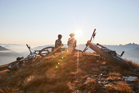 Zwei Mountainbiker stehen bei ihren Rädern auf einem Gipfel im warmen Morgenlicht.