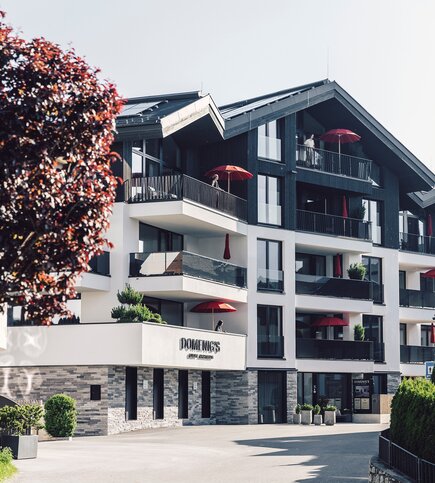 Außenansicht von Domenig's Luxury Apartments mit mehreren Balkonen und roten Sonnenschirmen.