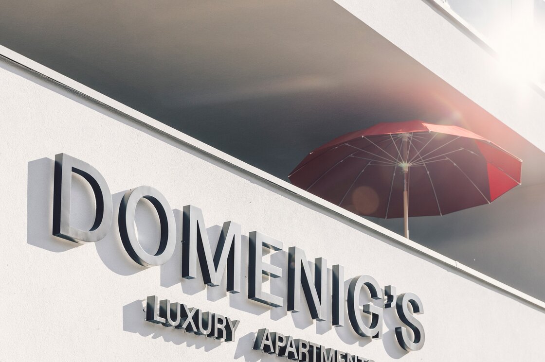 Das Schild von Domenig's Luxury Apartments an einer weißen Fassade mit rotem Sonnenschirm.