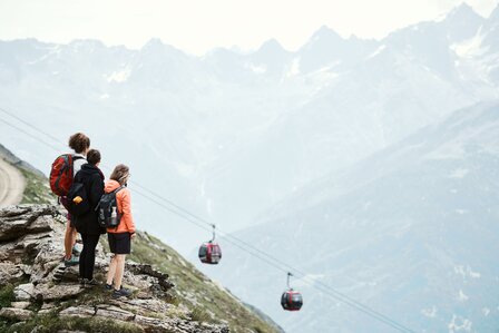 Drei Wanderer stehen auf einem Bergpfad und schauen auf die Seilbahn über dem Tal.