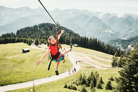 Eine Frau fährt an einer Sommerrodelbahn über eine grüne Bergwiese mit Blick auf die Alpen.