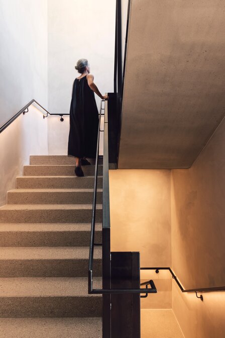 Eine Frau in dunklem Kleid geht eine Treppe in einem modern gestalteten Treppenhaus hinauf.