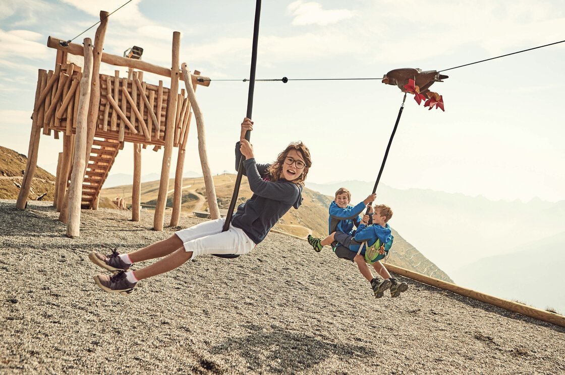 Drei Kinder spielen auf einer Seilbahn am Bergspielplatz mit Holzkonstruktion.