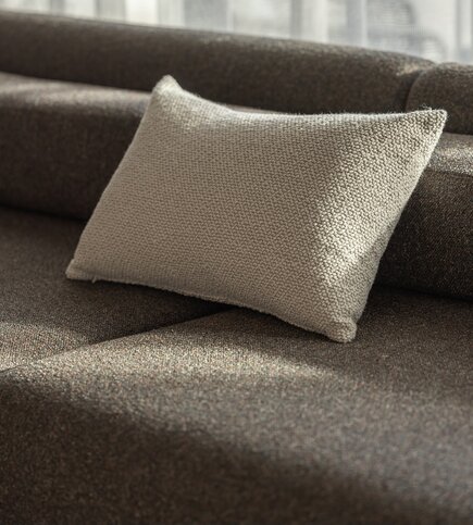 Ein helles Textilkissen liegt auf einem braunen Sofa im weichen Tageslicht.
