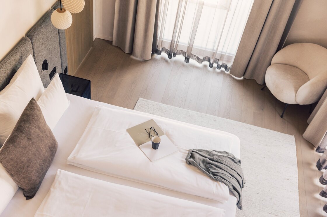 Ein modernes Doppelzimmer mit zwei Betten, hellen Vorhängen und einem gemütlichen Sessel am Fenster.