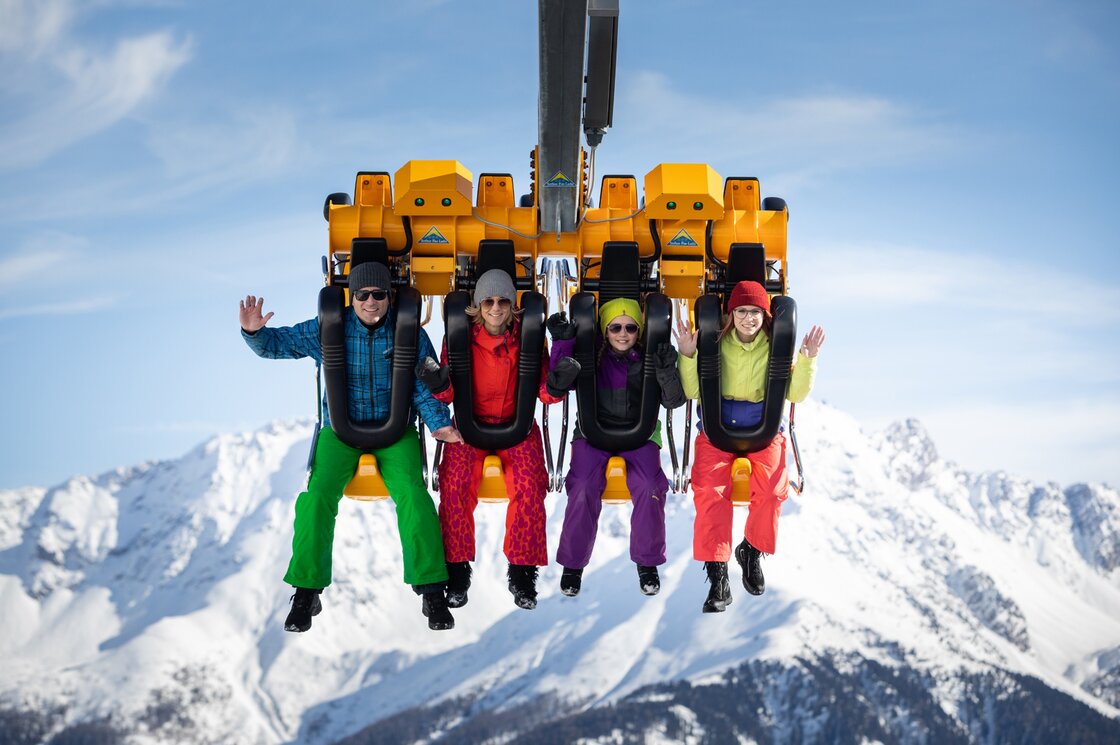 Vier Wintersportler sitzen in einem Sessellift vor verschneiten Bergen und winken in die Kamera.