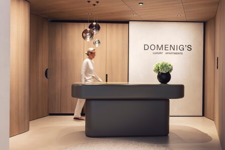 Ein moderner Empfangsbereich mit dunklem Tresen, Holzverkleidung und Domenig's Luxury Apartments im Hintergrund.