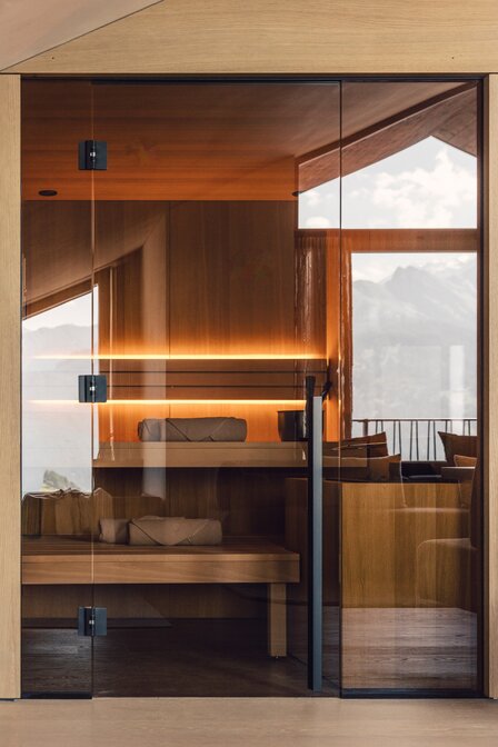 Eine moderne Sauna mit Glasfront und Blick auf die Berge sowie Sitzbank im warmen Holzinterieur.