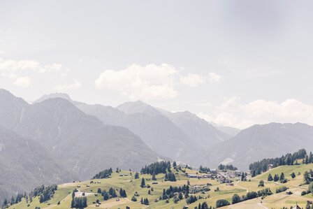 Weite Berglandschaft mit grünen Hängen und fernen Gipfeln unter hellem Himmel.