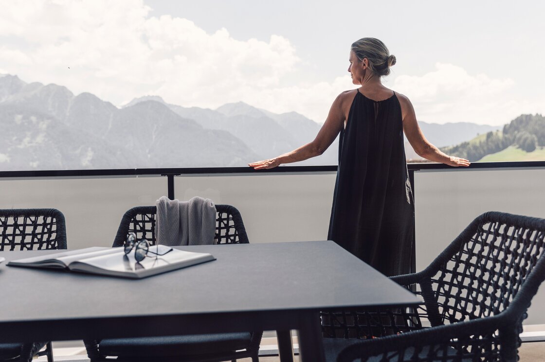 Eine Frau steht auf dem Balkon und blickt auf die Berge, vor ihr stehen ein Tisch und Stühle.