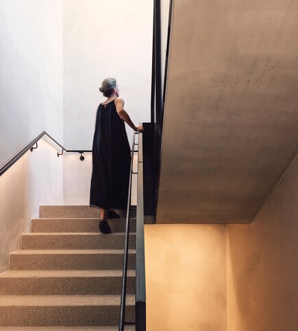 Eine Frau geht eine schmale Treppe mit schwarzem Geländer in einem modernen Innenraum hinauf.