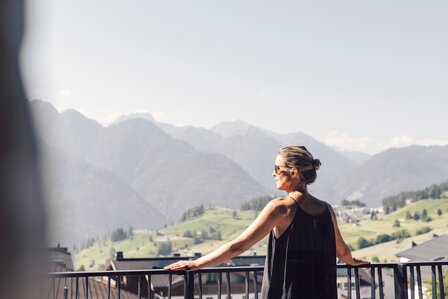 Eine Frau lehnt am Balkon und blickt über die Berge und Wiesen von Fiss.