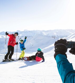 Skifahrer und eine snowboardende Person posieren auf einer verschneiten Piste vor den Bergen.