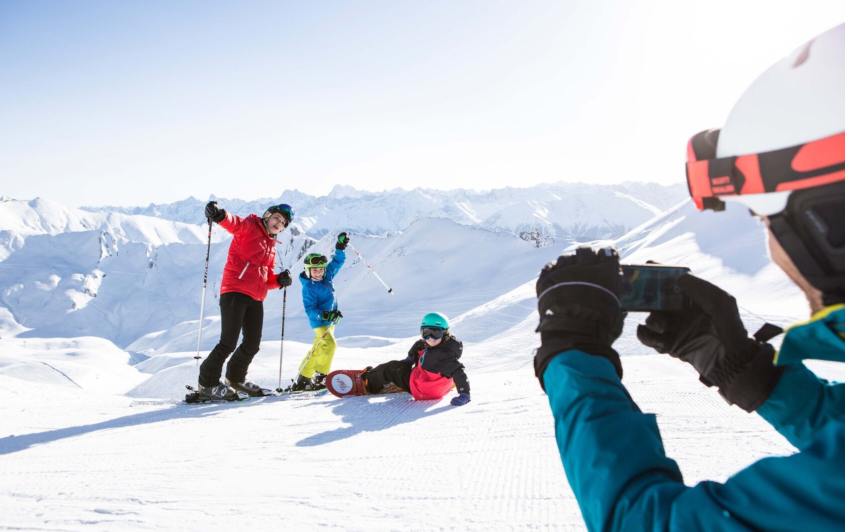 Skifahrer und eine snowboardende Person posieren auf einer verschneiten Piste vor den Bergen.