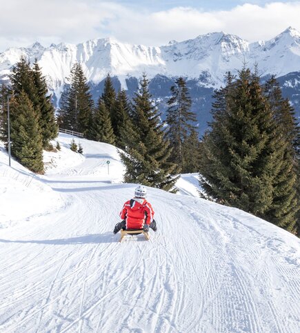 Ein Kind fährt auf einem Schlitten eine präparierte Winterpiste hinunter, umgeben von verschneiten Tannen und Bergen.