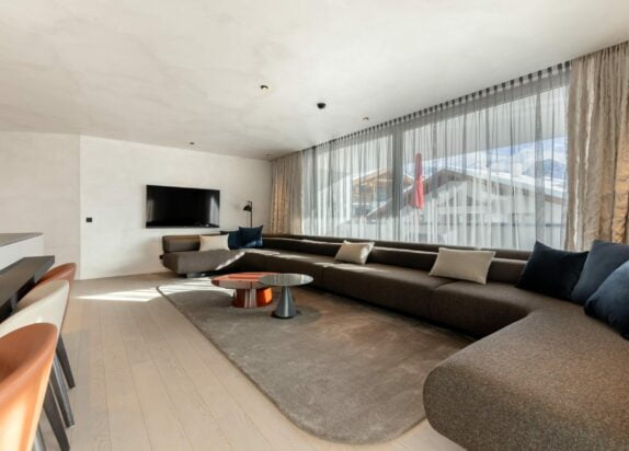 Übersicht Wohnbereich mit großer grauer Couch, Kissen, großes Fenster, TV, grauer Teppich