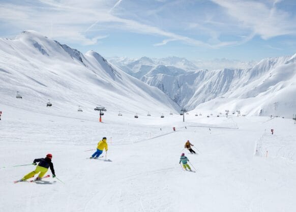 Skigebiet für Familien, Familie fährt Ski auf einer breiten weißen Skipiste, links ist ein Sessellift