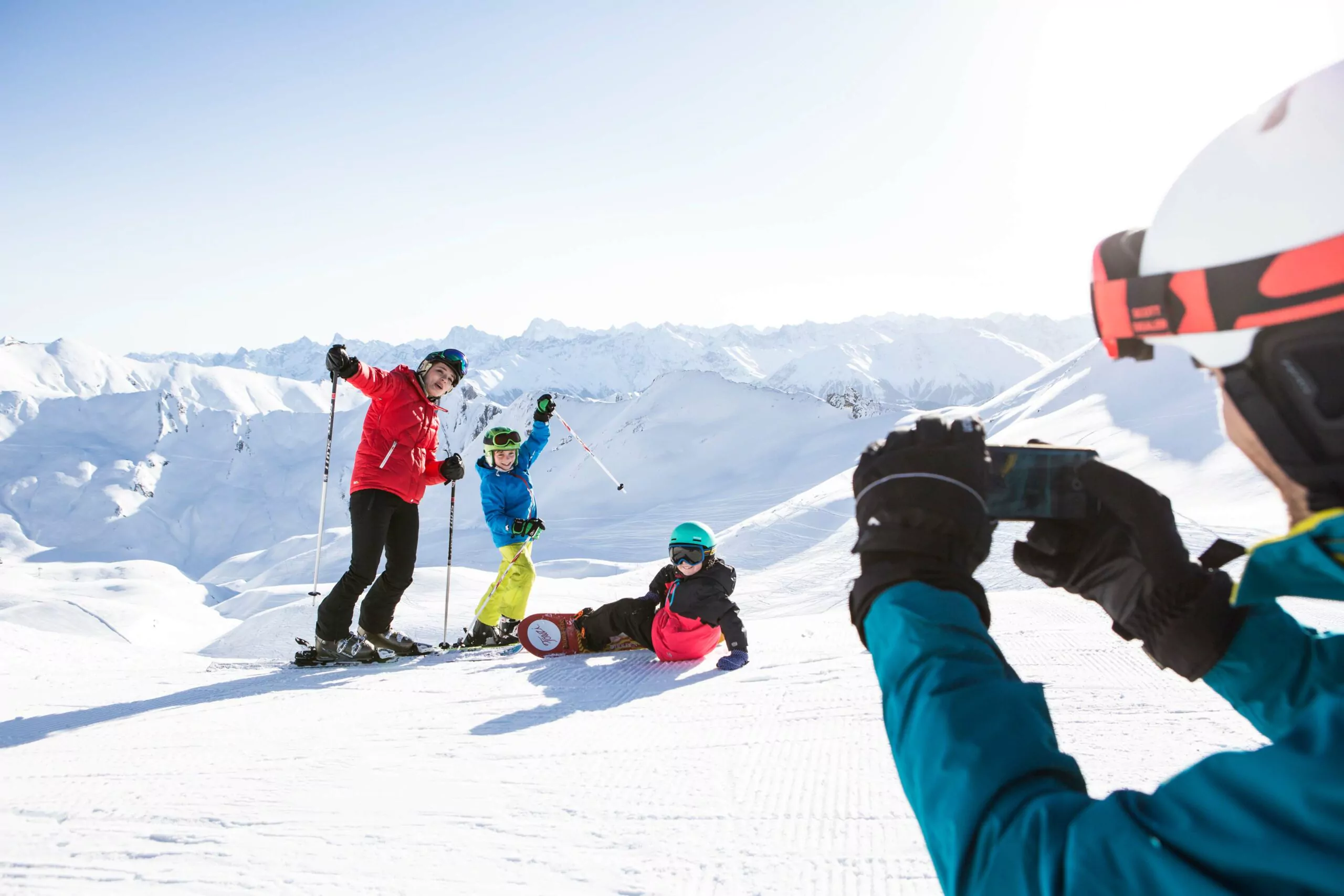 Skiurlaub mit Kindern, Papa macht Foto von Mama und zwei Kindern, Berge und Schnee im Hintergrund