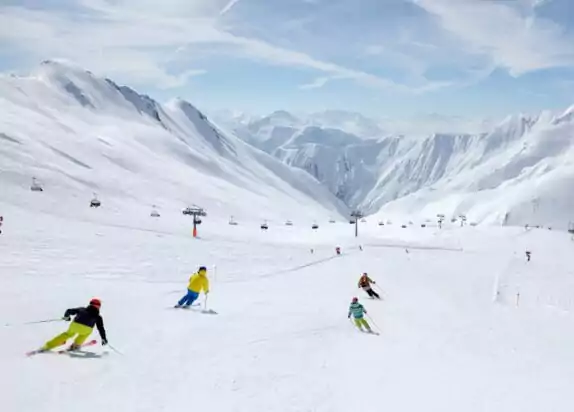 Skigebiet für Familien, Familie fährt Ski auf einer breiten weißen Skipiste, links ist ein Sessellift